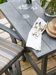 Outdoor wooden dining set with a gray table, striped cushioned chairs, and a place setting of napkin, forks, spoons, and plates on the table.