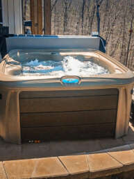 Outdoor hot tub bubbling on a stone patio beside a house, with a forested yard in the background.