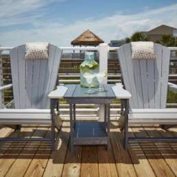 Two gray Adirondack chairs with cushions on a wooden deck, a small table between them holding a pitcher and two glasses, ocean view in the background.