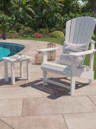 White Adirondack chair with a light cushion beside a blue pool on a beige tiled patio, with decorative lanterns nearby.