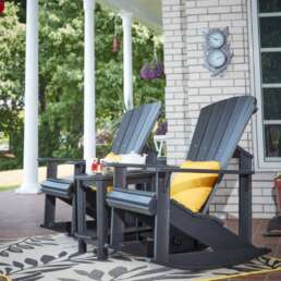Two dark wooden rocking chairs with bright yellow cushions sit on a patterned outdoor rug on a brick porch, with a coffee table holding a teapot and mugs nearby.
