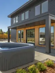 Modern backyard with a gray hot tub on a paved patio beside a two‑story house with wooden accent walls in a contemporary setting.