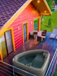 Red cabin with yellow trim, a wooden deck, and a large hot tub next to outdoor seating. A green lawn stretches beyond the railing.
