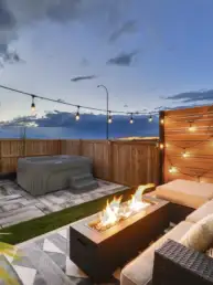 Cozy backyard patio with string lights, a rectangular fire pit, and cushioned seating under a wooden privacy fence at dusk/early evening.