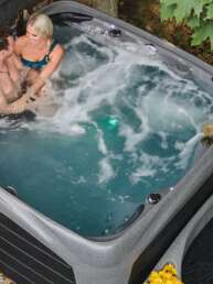 Couple relaxing in a gray outdoor hot tub on a patio with yellow flowers nearby and leaves on the ground.