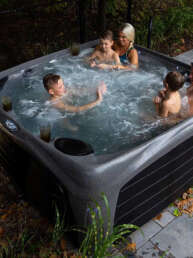 Family of five enjoying a soak in an outdoor hot tub on a patio, autumn leaves on the ground nearby.