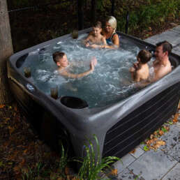 Family of five enjoying a soak in an outdoor hot tub on a patio, autumn leaves on the ground nearby.