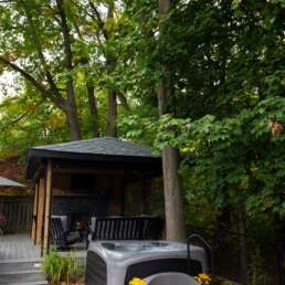 Backyard patio with a gray hot tub in the foreground, seating area under a wooden gazebo, surrounded by tall green trees.
