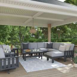 Outdoor covered patio with a grey sectional sofa, matching chairs, and a coffee table on a wooden deck surrounded by greenery, with a pendant light overhead.