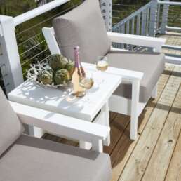 Outdoor white patio seating on a wooden deck with gray-cushioned chairs; a white table holds a pink wine bottle, two glasses, and a bowl of artichokes.