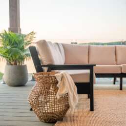 Outdoor lounge area with beige cushioned sofa on a wooden deck, a woven basket with a blanket, and a potted plant by the chair.