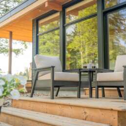 Outdoor wooden deck with two beige-cushioned chairs and a small table beside a glass-front house, amid green trees.