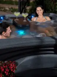 Three friends relaxing in an outdoor hot tub with drinks on a tray at night.