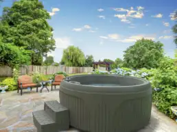 Round gray hot tub on a stone patio with two orange-cushioned chairs and a small table, surrounded by hydrangeas and trees under a blue sky.