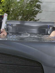 A man and a woman soak in a dark gray hot tub in a sunny backyard, each reading a book.