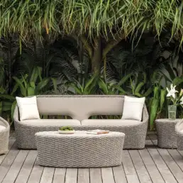 Outdoor wicker seating set on a wooden deck with beige cushions and white flowers in a vase, surrounded by tropical plants.