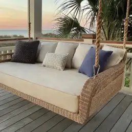 Wicker porch swing sofa with beige cushions and assorted throw pillows on a wooden deck at sunset by the coast.
