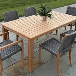 Outdoor wooden dining table with six gray woven chairs on a stone patio, a small plant centerpiece, and a grassy lawn in the background.