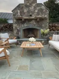 Stone outdoor fireplace with cushioned wooden seating around a wooden coffee table on a slate patio, surrounded by stacked firewood and greenery.