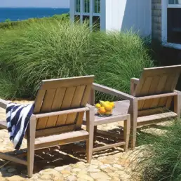 Two wooden outdoor chairs with a striped towel, a small table with a bowl of lemons, in a coastal stone patio by a house.