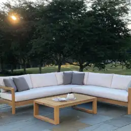 Outdoor wooden sectional sofa with white cushions and gray pillows on a stone patio, with a matching coffee table and a vase of flowers on a side table in a sunny backyard.