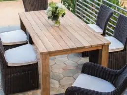 Outdoor wooden dining table with six wicker chairs and a vase of flowers on a stone patio. cushions are light gray.
