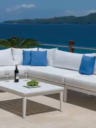 White outdoor sectional with blue cushions on a balcony overlooking the sea; coffee table holds a wine bottle, two glasses, and grapes awaiting.