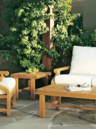 Outdoor wooden patio set with white cushioned chairs and sofa, a matching coffee table and side table on a stone patio surrounded by greenery, with a plate of lemons and magazines on the table
