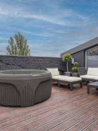 Rooftop patio with a circular hot tub, a beige-cushioned sofa set, and a wooden deck against a dark stone wall and blue sky.