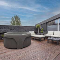 Rooftop patio with a circular hot tub, a beige-cushioned sofa set, and a wooden deck against a dark stone wall and blue sky.