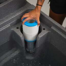 Hands install a white pleated pool filter cartridge with a blue end cap into a grey filter housing in a spa or hot tub.