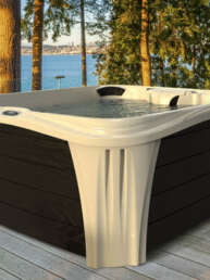 White and dark-paneled outdoor hot tub on a wooden deck by a lake, with trees and a distant shoreline in view.