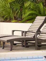 Two wooden poolside lounge chairs with matching ottomans beside a tiled pool edge and tropical plants in the background