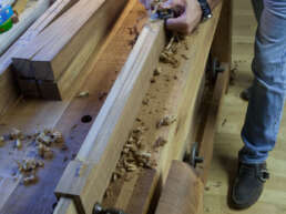 Person planing a long wooden beam on a workbench, with wood shavings scattered around and several chisels and tools nearby in a woodworking shop