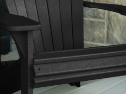Close-up of a dark outdoor bench with a wooden armrest and metal slats, set on a porch with a stone wall in the background.